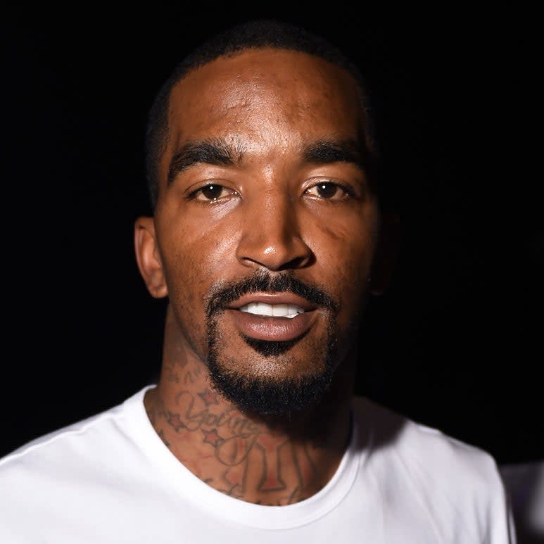 Man with tattoos on his neck and a goatee smiling, wearing a plain white T-shirt. No recognizable background