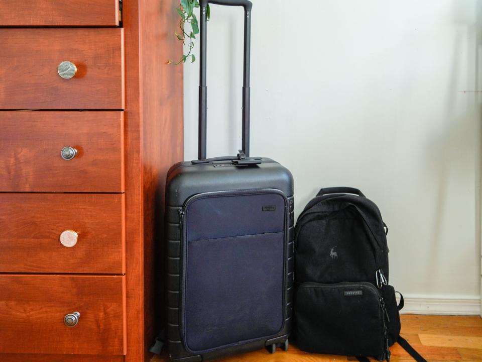 Black carry-on suitcase and backpack on a wood floor with a white wall behind and a wooden dresser on the left