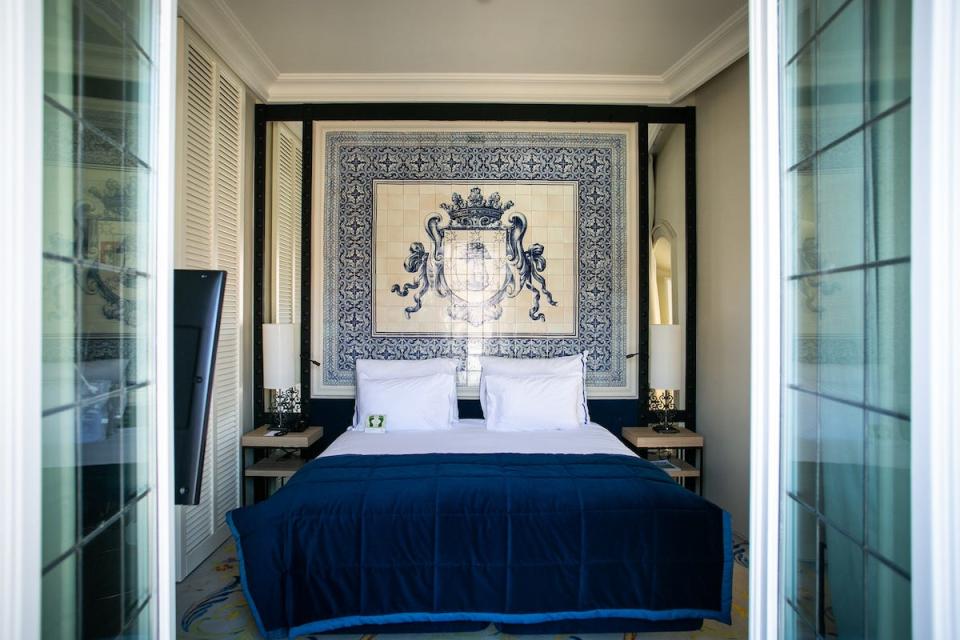 Doors open to a view of a double bed with a blue quilt and a tiled piece of art behind it in a luxury hotel.