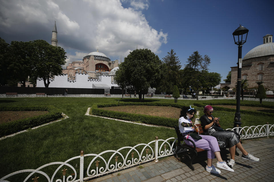 People sit in the plaza backdropped by a ''wall' set in front of Istanbul's 6th-century Hagia Sophia, the main cathedral of the Byzantine Empire which was converted into a mosque with the Ottoman conquest of the city, then known as Constantinople, in 1453, that was to be used as a set for the celebrations to mark the 567th anniversary of the conquest in Istanbul, Friday, May 29, 2020. A Muslim cleric recited the "prayer of conquest" from the Quran inside the Hagia Sophia. Muslim prayers at the UNESCO World Heritage site are highly controversial, hitting at the heart of the country's religious-secular divide. (AP Photo/Emrah Gurel)