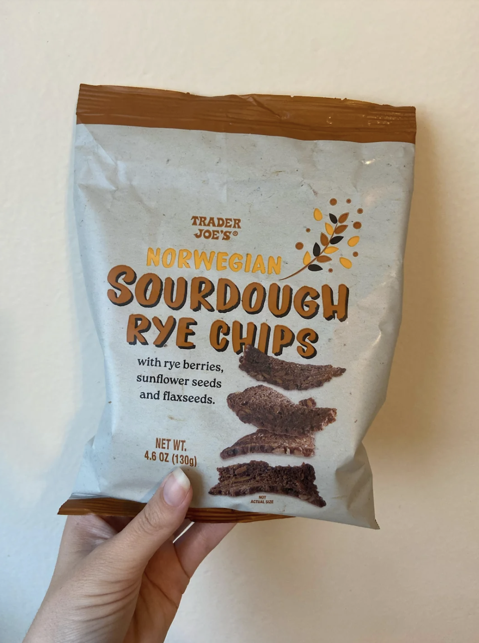 A hand holds a Trader Joe's Norwegian Sourdough Rye Chips bag. The chips contain rye berries, sunflower seeds, and flaxseeds. The bag weighs 4.6 oz (130g)