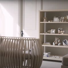 A beige rounded crib sitting in the middle of a neutral colored room with toys on a large bookshelf