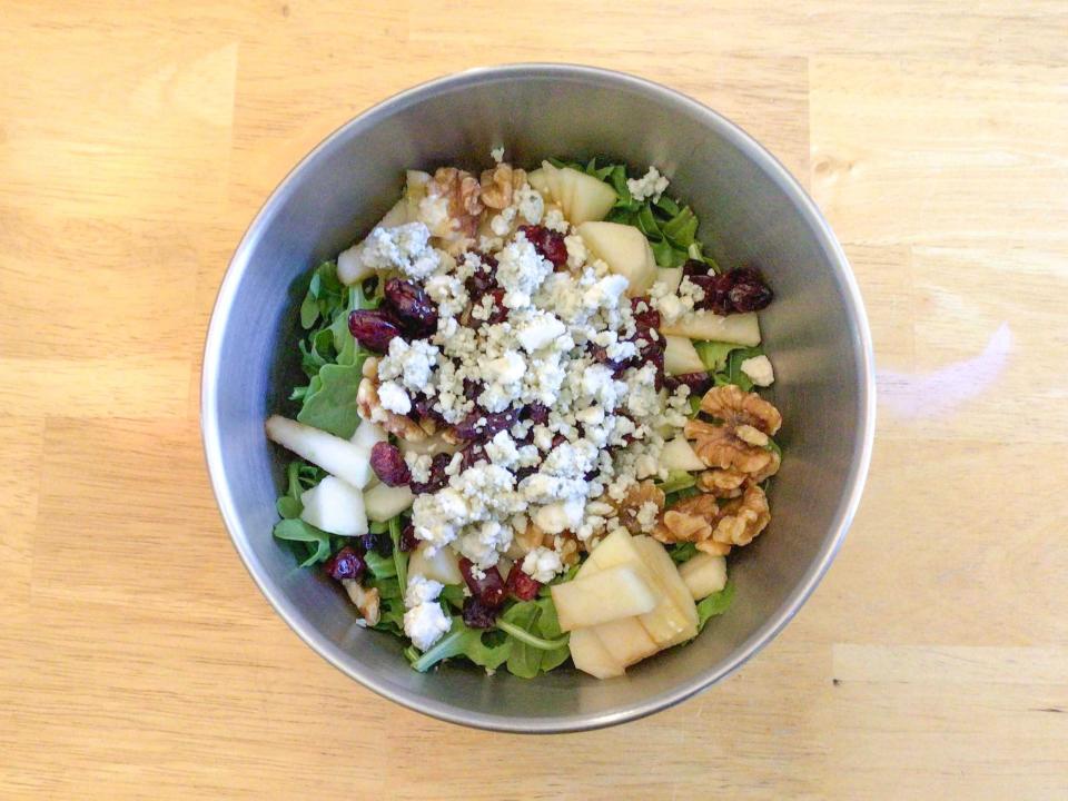 Bird's-eye-view of a metal bowl filled with arugula, nuts, cranberries, pieces of apple, and blue cheese on a wooden table