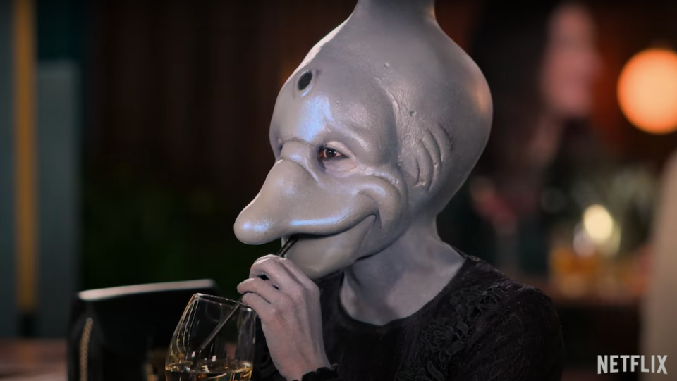 A person wearing dolphin prosthetics sips a cocktail from a straw.