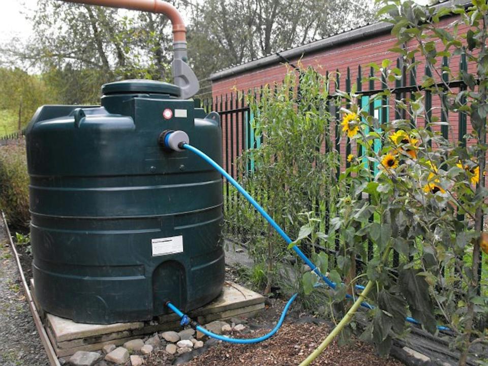A large tank with an inlet pipe above and pipes running from the bottom sits on a cement slab in a garden next to a fence with wildflowers lining it.