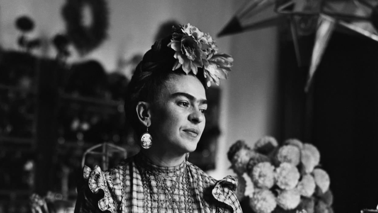 frida kahlo sits on a table while wearing a floral head piece, large earrings, a plaid blouse and striped pants, she looks off to the right