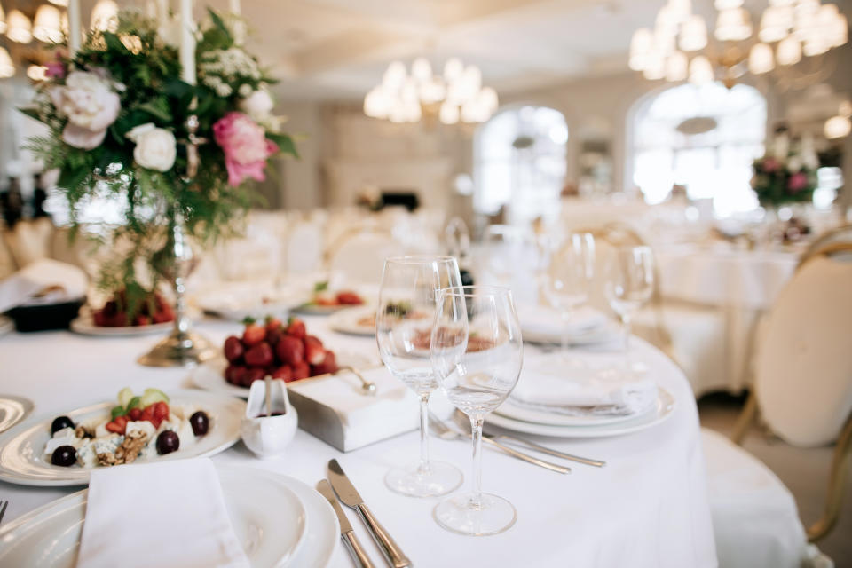 Elegant wedding reception setting with white linens, fine china, and elaborate floral centerpiece. Tables feature fruit, candles, and neatly arranged tableware