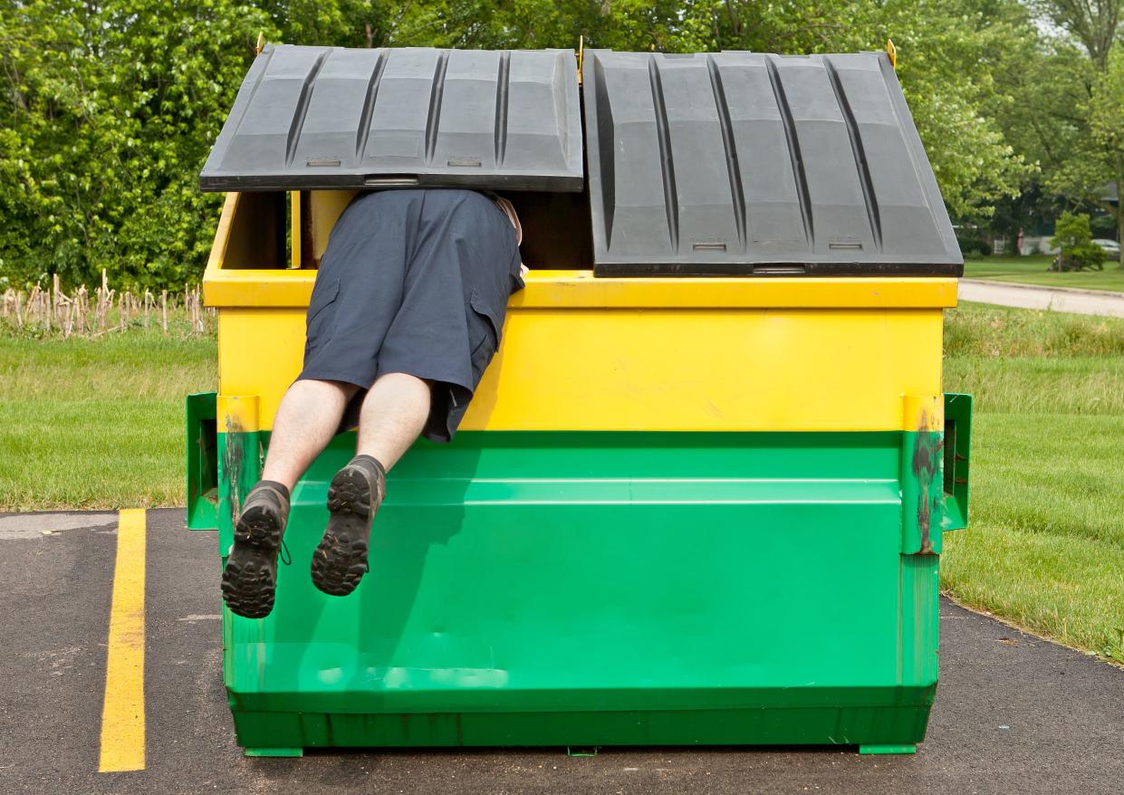 the legs and rear end of a caucasian man wearing shorts hangs out of a yellow and green dumpster