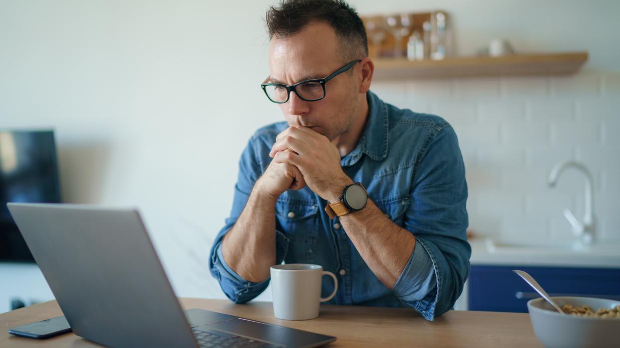  Man sitting at a table and looking at a laptop, holding one hand in the other in front of his face and looking concerned. 