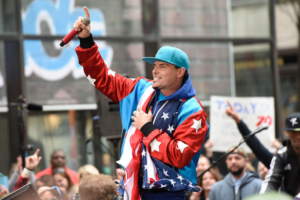 Vanilla Ice performs on an outdoor stage, holding a microphone and wearing a patriotic jacket and a baseball cap. He raises one arm, gesturing to the crowd