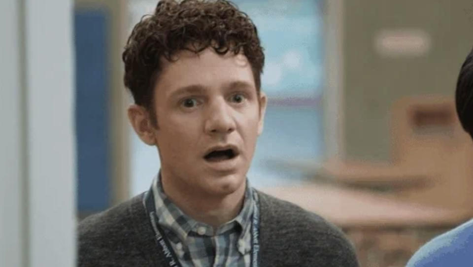 A man with curly hair and a surprised expression in a casual setting, wearing a checked shirt and cardigan