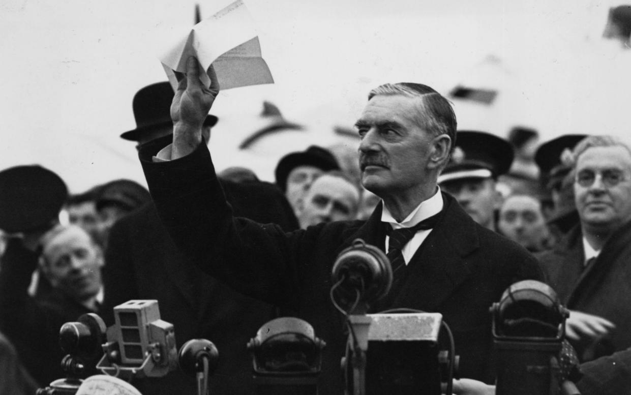 Chamberlain pictured in black and white, holding up the agreement signed by Adolf Hitler, with microphones ranged in front of him and press people surrounding him
