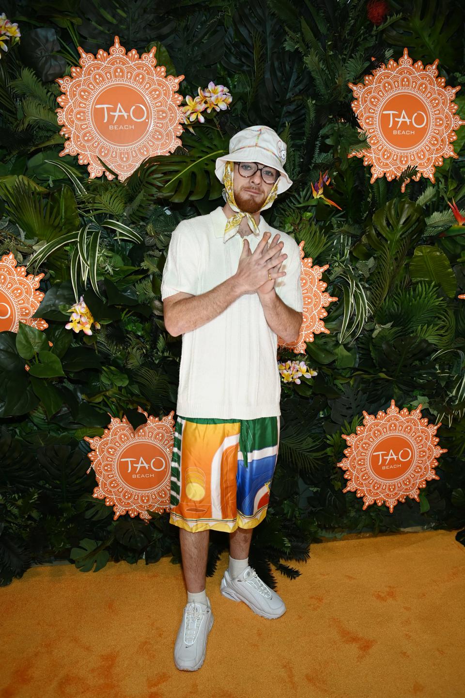 Angus in a white polo top and colorful printed basketball shorts. He's has a handkerchief tied around his head under a bucket hat.