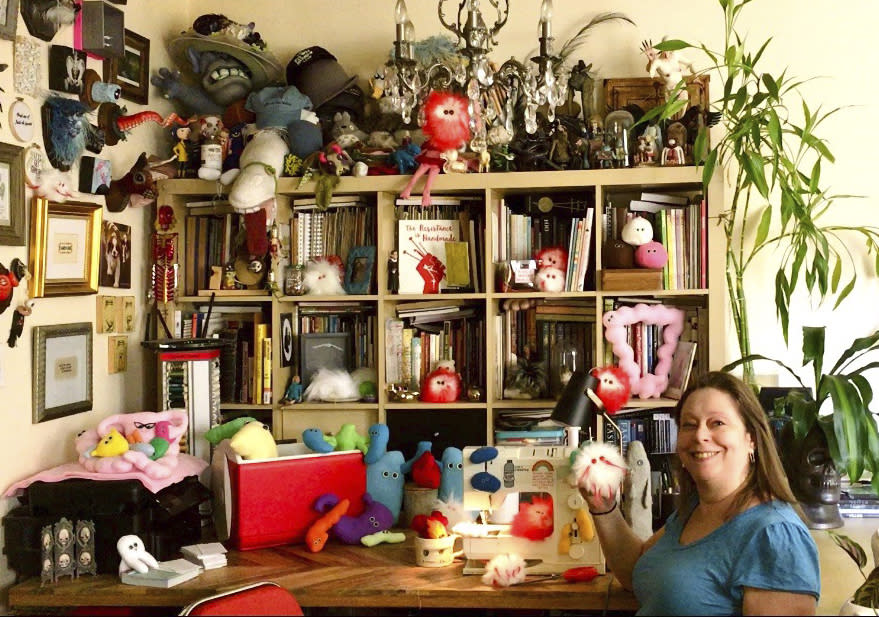 This July 25, 2020 photo shows Amy Micallef posing with her craft creations at her home. While Broadway stages remain dark due to the pandemic, Broadway workers are concentrating on side hustles. Micallef, a Broadway seamstress who has worked in the wardrobe departments of “Hamilton,” “Waitress” and “Frozen,” makes gleeful representations of COVID-19 for sale, complete with a pair of eyes and faux fur. (Amy Micallef via AP)