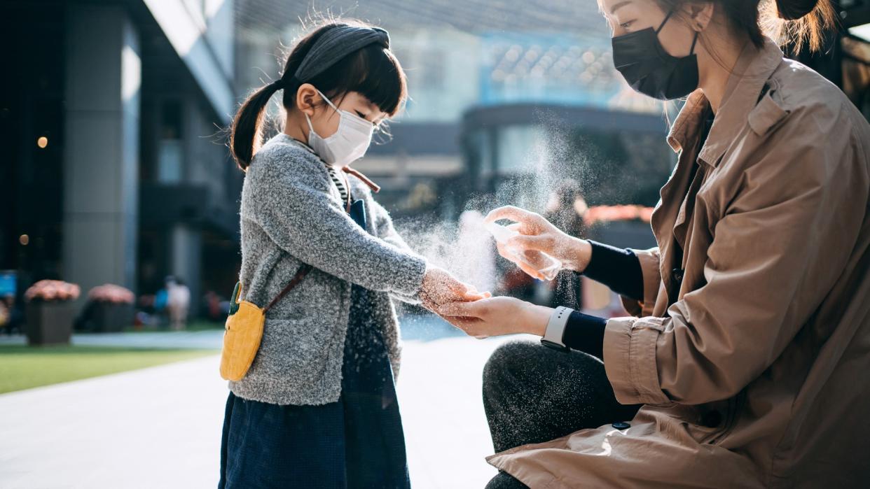 Mother applies hand sanitizer to her young daughter outdoors. Both are wearing protective face masks