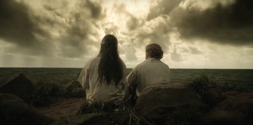 Two characters sit on rocks by the sea looking at a stormy sky in a scene from the show "Our Flag Means Death."