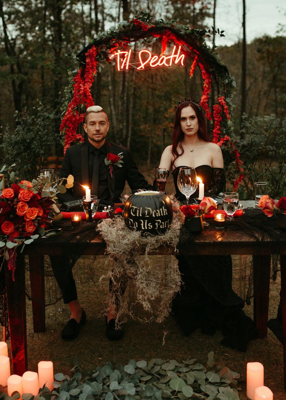 A bride and groom sit at a table in front of an archway that reads "Til Death" on it.