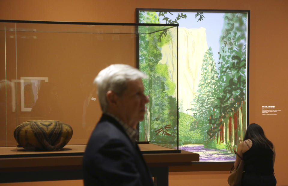 In this Wednesday, Oct. 23, 2019, photo, Richard Benefield, former executive director of the David Hockney Foundation, center, looks inside the exhibit of Hockney's Yosemite artistic work, background, along with baskets from weavers from the Miwok and Mono Lake Paiute tribes on display at the Heard Museum, in Phoenix. "David Hockney's Yosemite and Masters of California Basketry" exhibition opens Monday. (AP Photo/Ross D. Franklin)