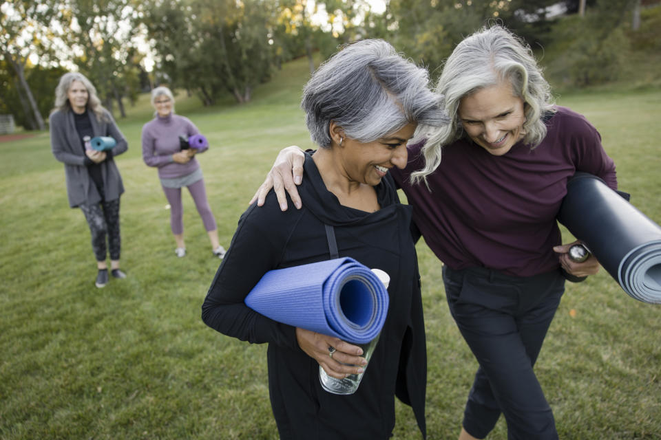 Happy mature women friends hugging, walking and laughing with yoga mats in park pension