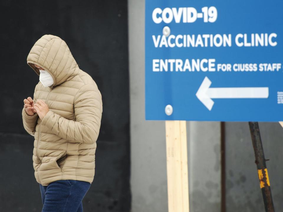 A person wearing a hooded coat and a face masks walks past a sign pointing to the entrance of COVID-19 vaccination clinic.