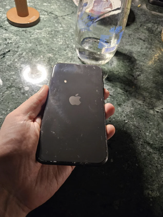 A hand holding a black Apple iPhone with a visible Apple logo on the back. A water bottle and other objects are on the table in the background