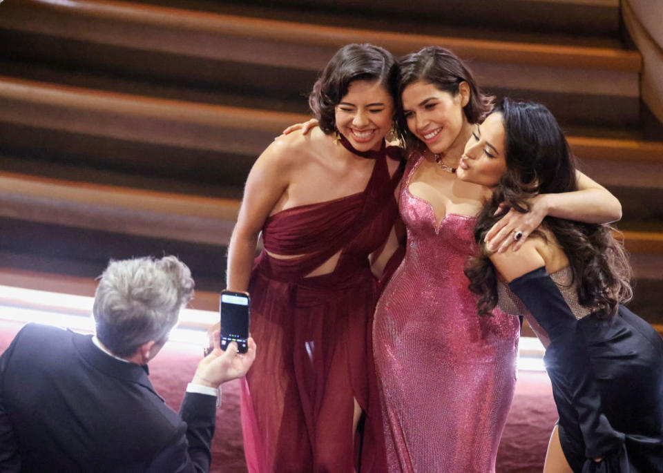 Three women hugging and posing for a selfie at a glamourous event, with a man taking the photo