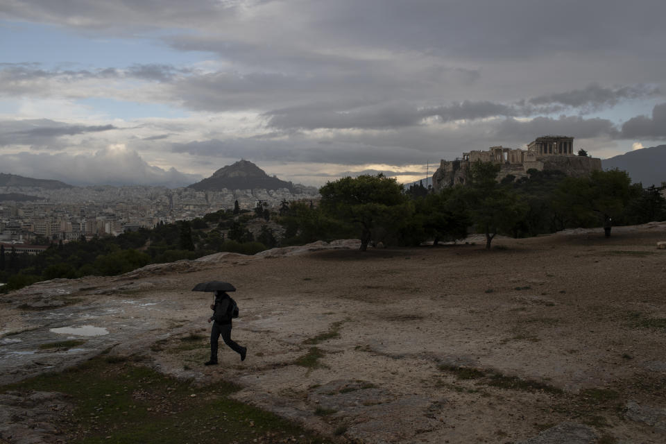 A man with an umbrella walks in the rain on Pnyx hill in front the ancient Acropolis hill, with the ruins of the fifth century BC Parthenon temple, in Athens, on Monday, Nov. 30, 2020 Greek officials say the number of new infections is waning in most parts of the country, which has been in lockdown for weeks.(AP Photo/Petros Giannakouris)