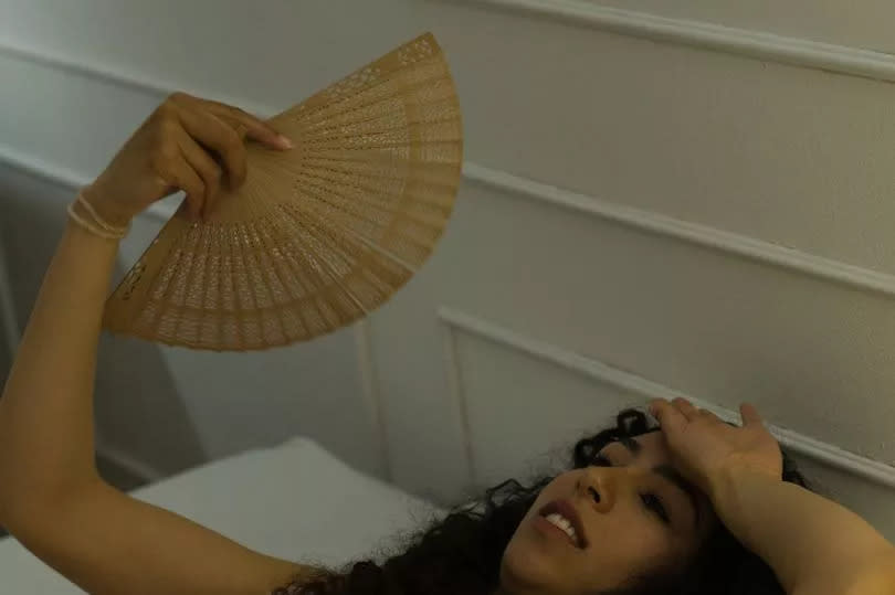 Upset young woman in pajamas feeling hot and using a fan during the summer in bed and having sleeping problems