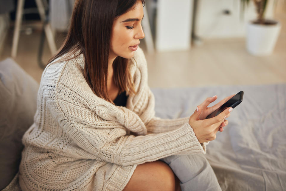 Attractive caucasian brunette dressed in beige sweater sitting on bed in bedroom and using smart phone.