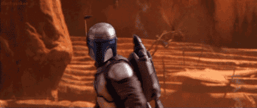 Character in Mandalorian armor walking in a desert-like setting from the TV series "The Mandalorian."