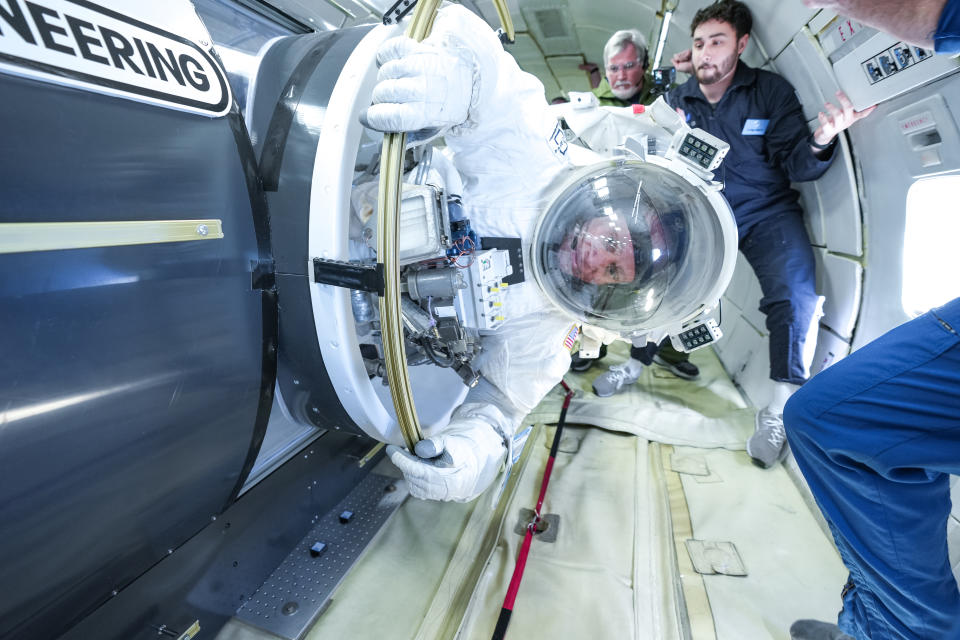 a person in a bulky spacesuit floats in an aircraft fuselage surrounded by people in blue flight suits