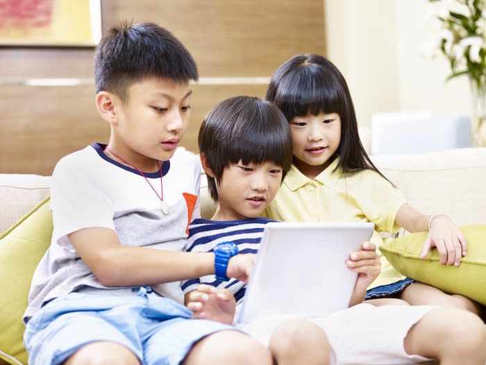 Three young children sitting on a couch looking at a tablet.