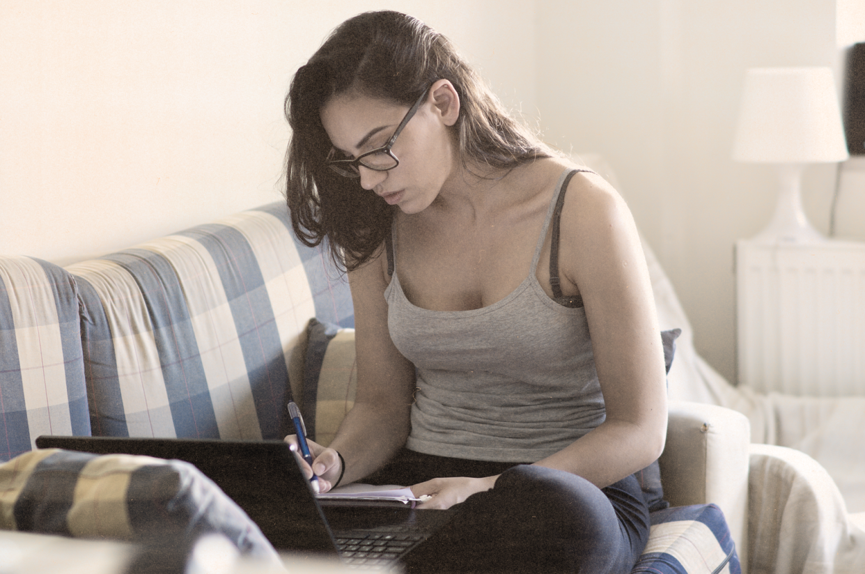 Person sitting on a couch writing in a notebook in front of a laptop