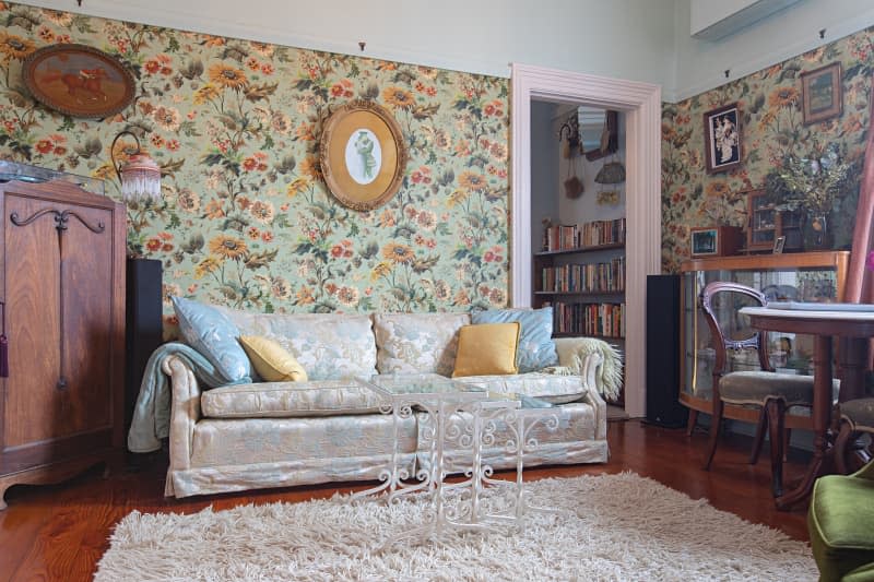 A living room with floral wallpaper, floral couch and fuzzy rug.