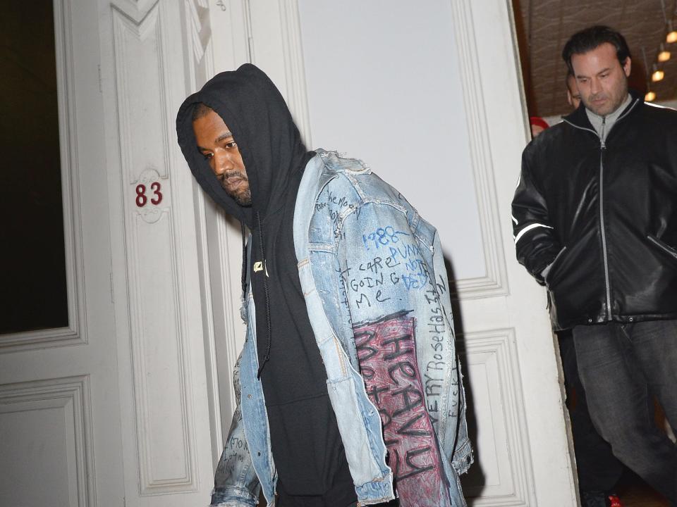 kanye west stepping out onto metal steps outside a building in soho, wearing a jean jacket with pen writing on it and a black hoodie. behind him, coming out of the door, is security detail steve stanulis, a man wearing a black leather jacket with white detailing, and slicked back hair