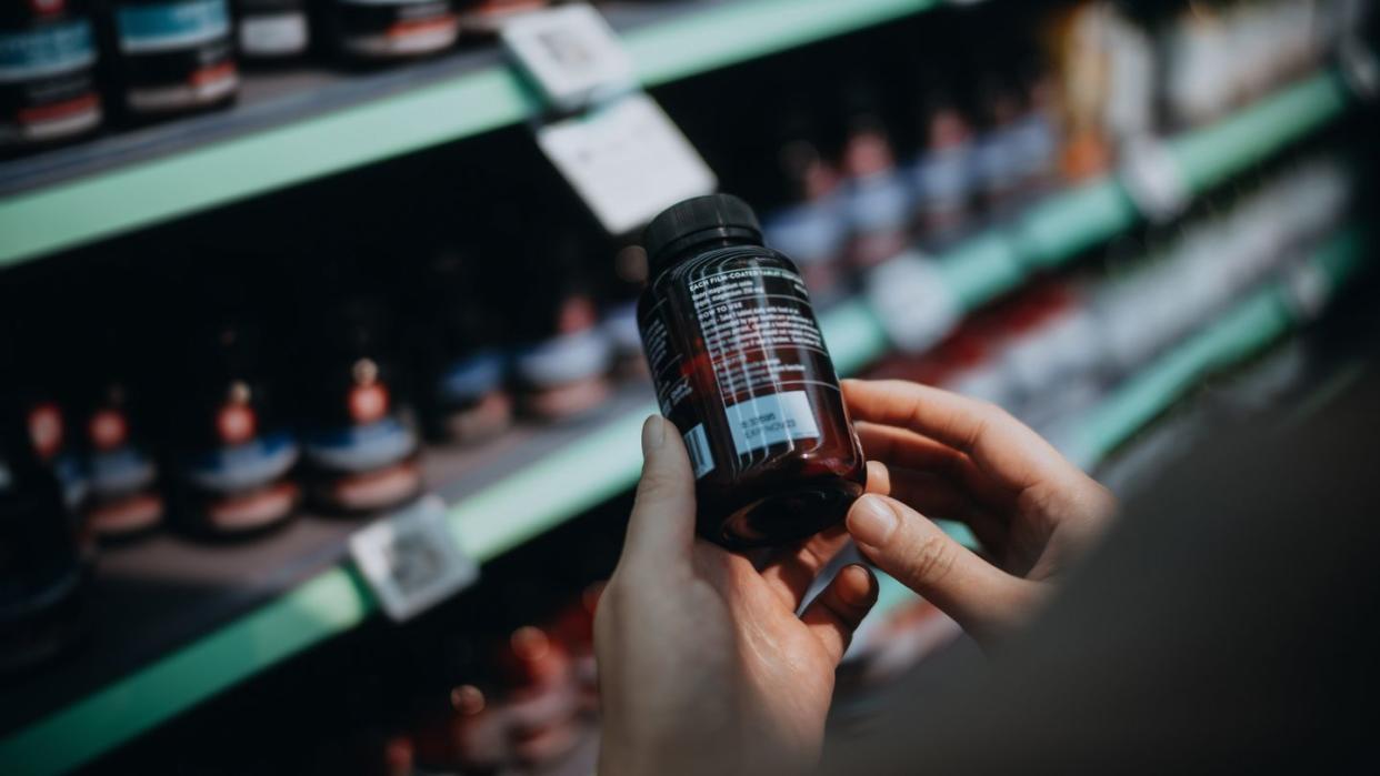 over the shoulder view of woman browsing through medical products and reading the label on a bottle of medicine in front of the shelves in a pharmacy healthcare, medication and people concept