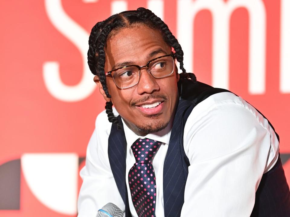 nick cannon sitting onstage, holding a microphone and wearing a vest and tie as he looks to the side