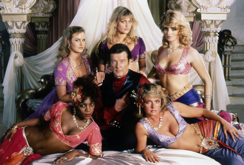 Actor Roger Moore surrounded by women on bed on set of film