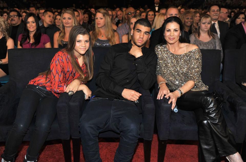 miles richie and diane alexander pose for a photo while sitting in a crowded theater, miles wears and all black outfit and lefts one hand to his chine, diane wears a sparkly gold and silver top with black pants and smiles