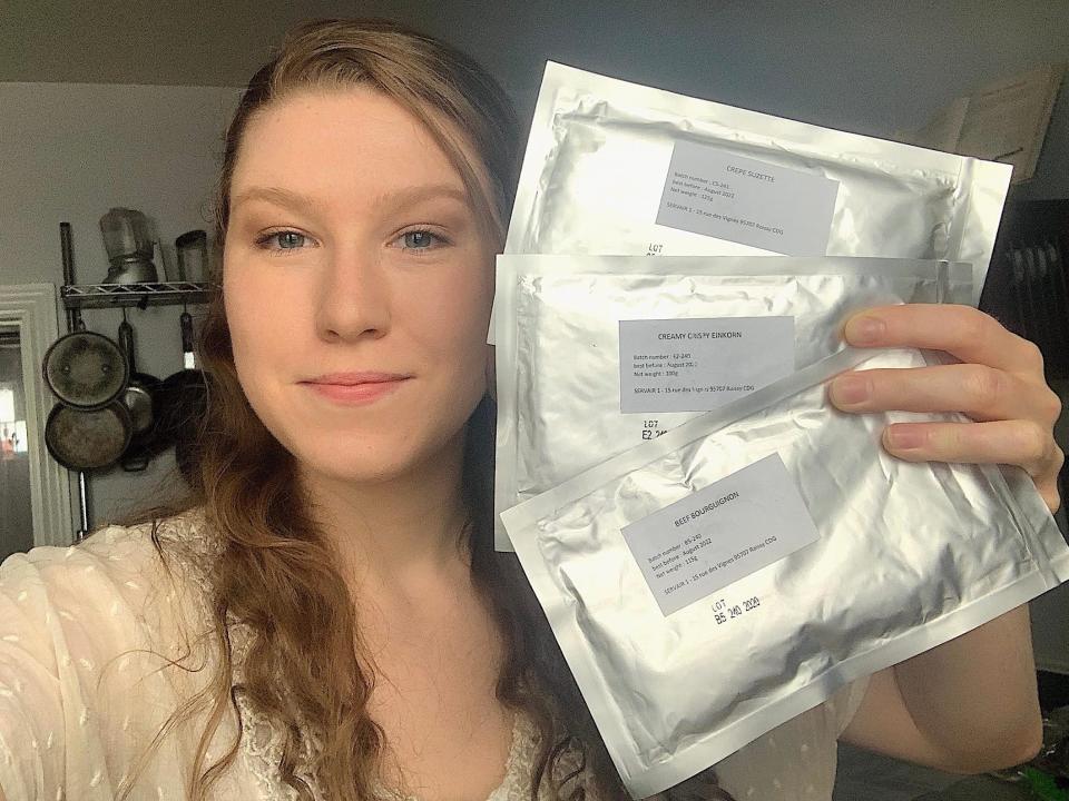 smiling woman holds up three shiny foil packets of astronaut food in kitchen