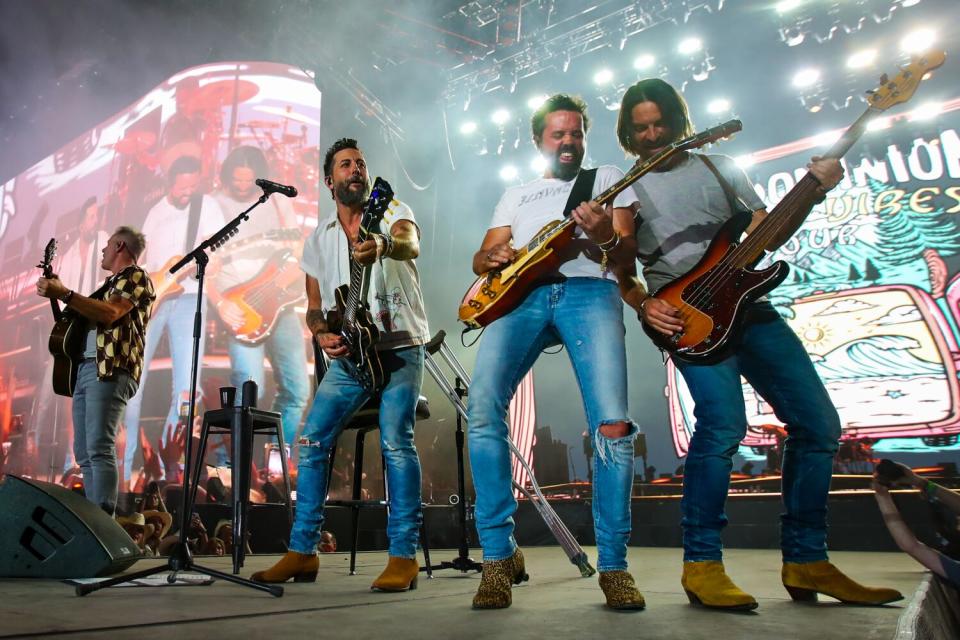 Four men play various kinds of guitars on stage while a huge screen behind them shows them playing.