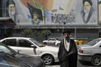 In this Friday, May 17, 2019 photo, a cleric crosses Enqelab-e-Eslami (Islamic Revolution) street in front of a mural depicting late Iranian revolutionary founder Ayatollah Khomeini, right, Supreme Leader Ayatollah Ali Khamenei, left, and the Basij paramilitary force, in downtown Tehran, Iran. (AP Photo/Vahid Salemi)