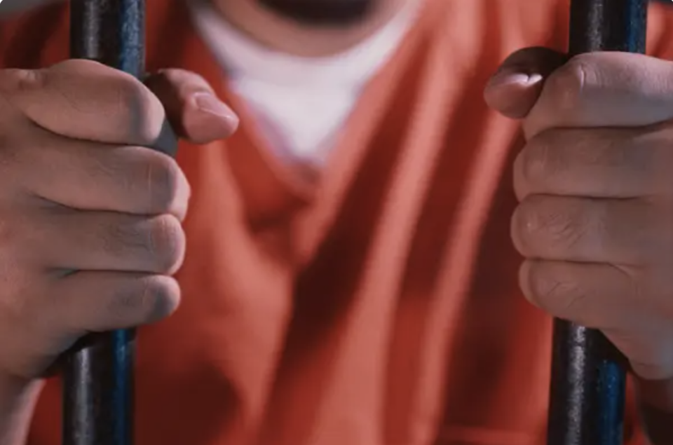 A person in an orange prison uniform holds on to jail cell bars with both hands