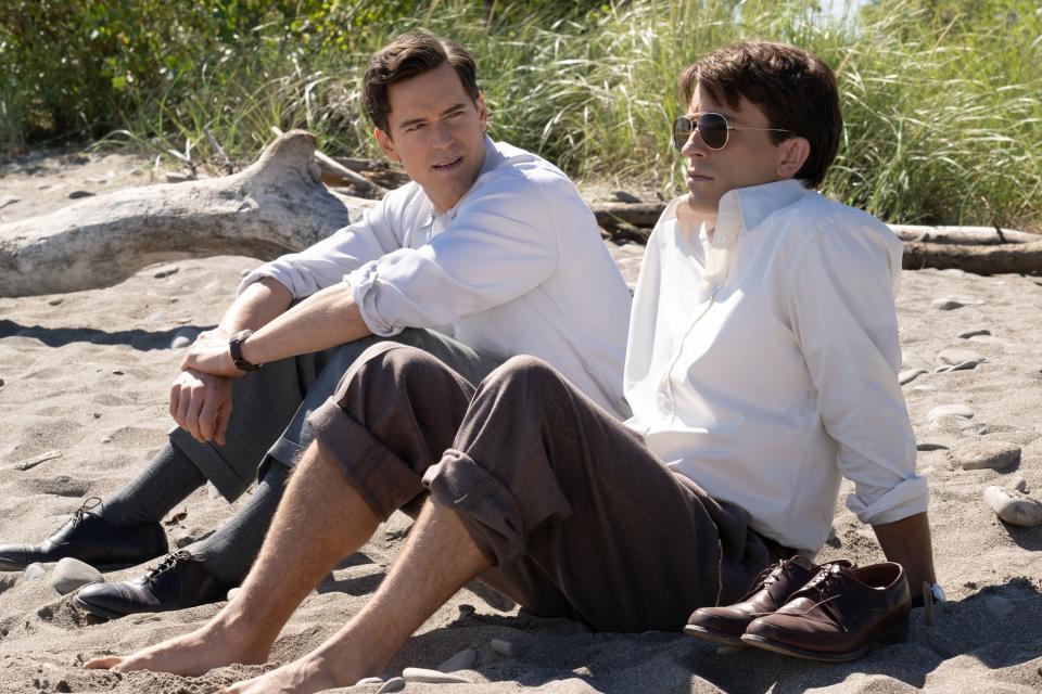 Matthew Bomer and Jonathan Bailey sit on a sandy beach, casually dressed in white shirts and pants, with shoes off