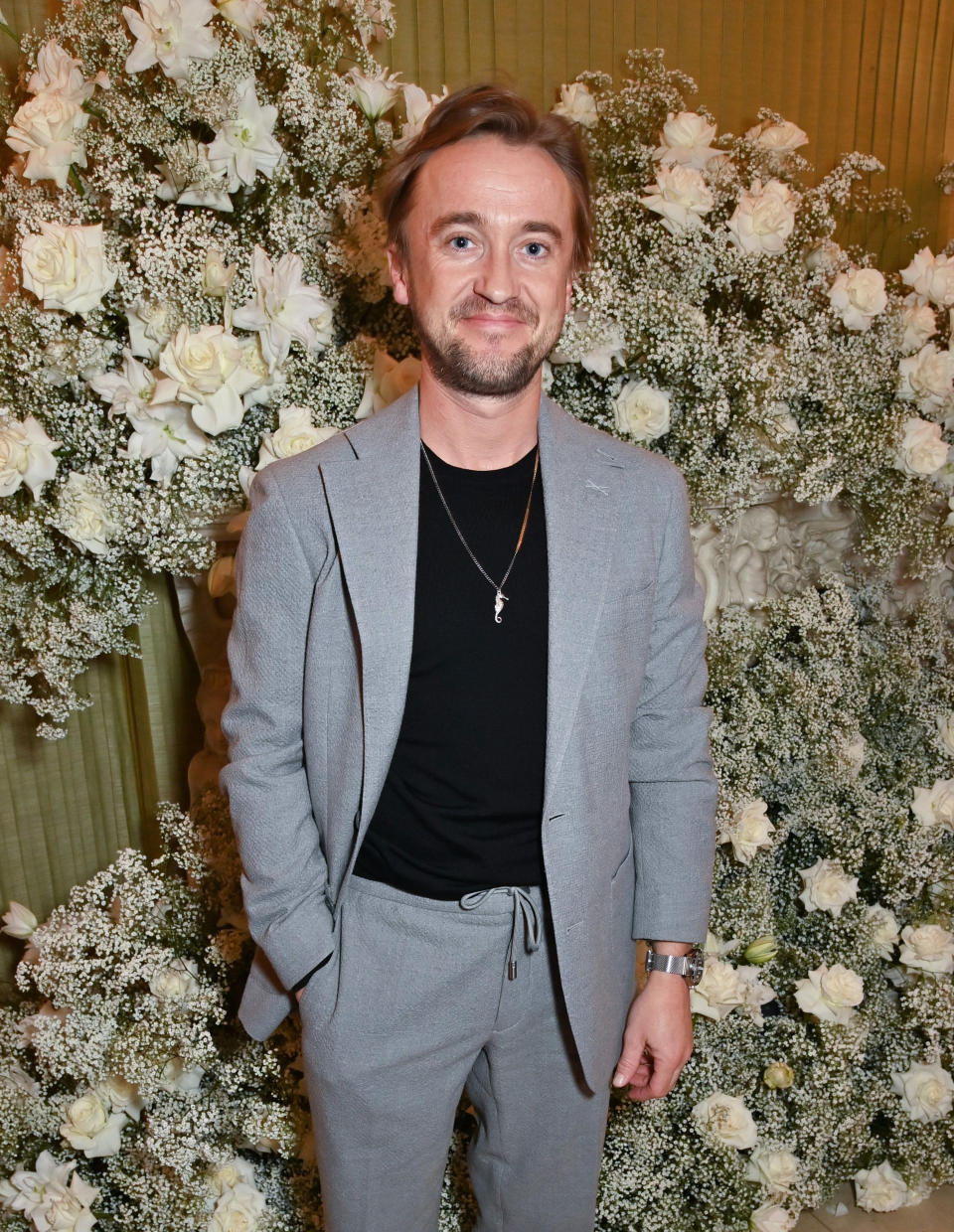 Tom Felton in a grey suit and black shirt, smiling, standing in front of a floral backdrop at an event