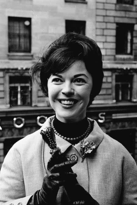 Shirley Temple smiles while posing outside an urban building, wearing a coat with a brooch, gloves, and a pearl necklace