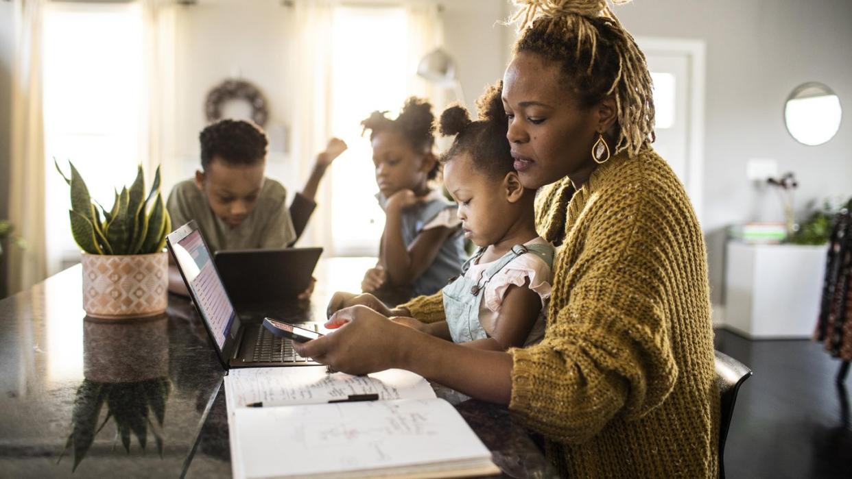 mother working from home while holding toddler, family in background