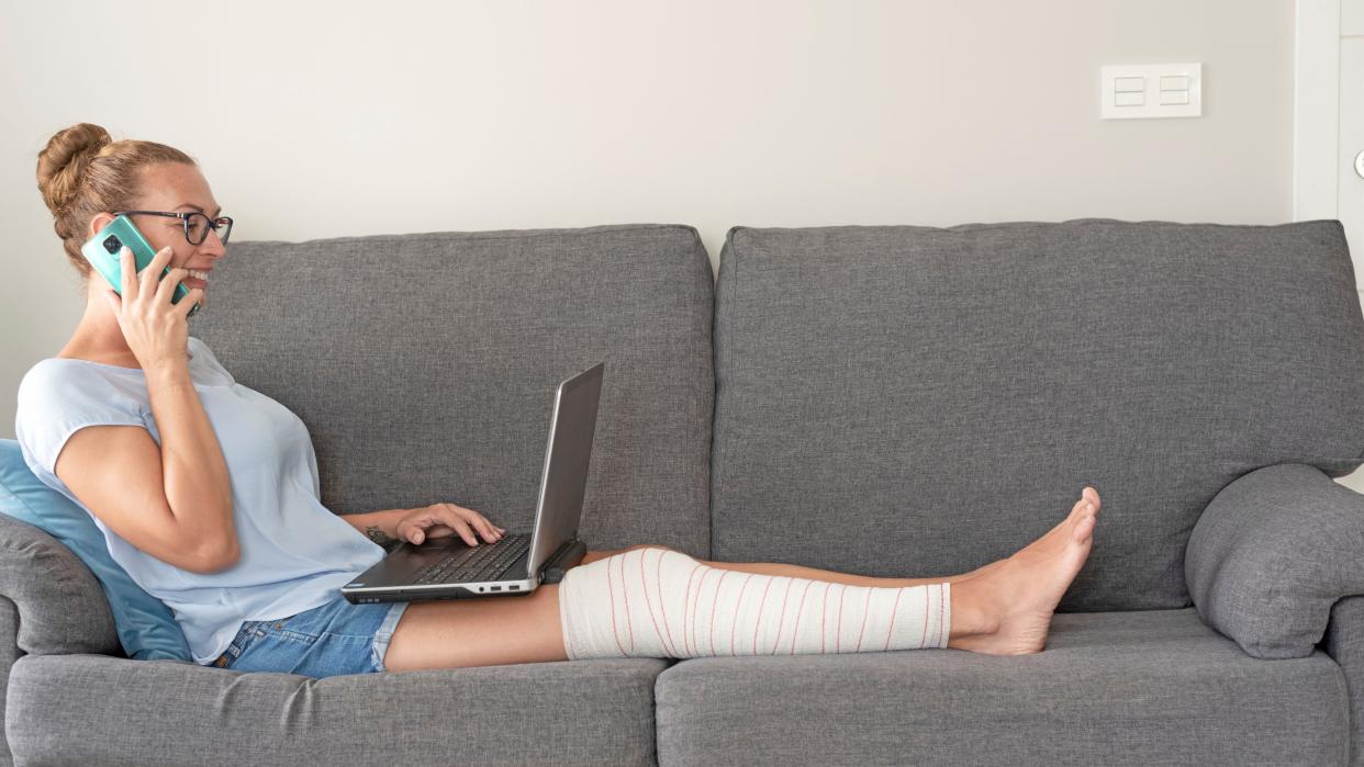  A young woman sitting on the sofa with her leg in bandage talking on the mobile phone and using the laptop 