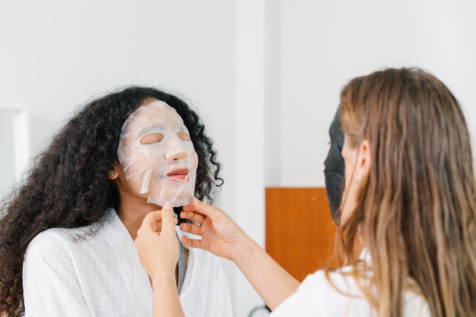 Two females putting on skincare masks for each other. One in a white, the other in a black mask.