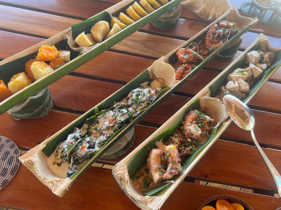 Fish, root vegetables, and other seafood cooked and served in pieces of halved bamboo with a spoon on the side of one plating arrangement
